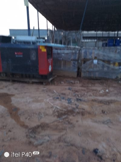 A muddy ground area is visible, with a portable generator on a wooden pallet to the left. Adjacent to the generator are large, possibly crated items, wrapped and secured with straps. A warehouse structure with a dark roof and supporting beams is in the background, partially obscured by the stacked materials. There is some visible structure in the rear. The bottom of the image has the text "itel P40 4G" in white.