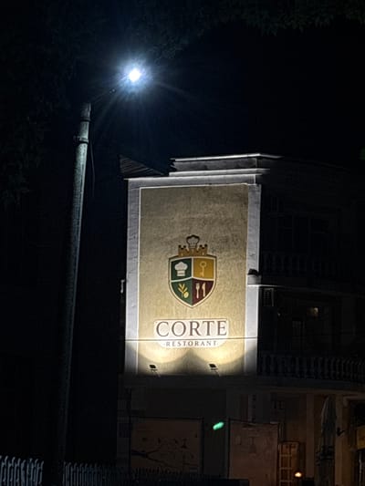 A night-time outdoor shot captures the illuminated sign of "CORTE RESTURANT" in Razgrad, Bulgaria. The sign is centered on a building facade, featuring a crest with culinary symbols such as a chef's hat, a key, and utensils, set against a textured golden background. The name "CORTE" is prominently displayed in large, capitalized letters above the word "RESTORANT." The scene is lit by a nearby streetlamp, casting a bright glare, and spotlights directed at the sign. The surrounding environment is dark, with the silhouette of trees and a white fence visible in the foreground. The building itself appears to have an upper floor with a balcony and windows, also dimly lit.