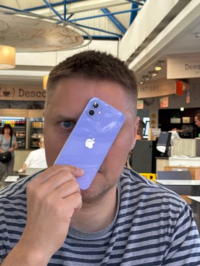 The image shows a man holding a purple iPhone to his ear. He is wearing a grey and black striped t-shirt. The background is blurred, but appears to be an indoor setting with shelves of products and other people. Some text is visible in the background, including "( ) Desc" and "ANT CAFE". The lighting suggests it is daytime. The man's eye is visible and appears to be looking towards the camera. The overall scene is casual, possibly a store or cafe.