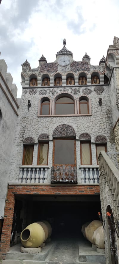 This is a low-angle, full-shot, exterior, eye-level photograph of a building in the city of Voskevaz, Armenia. The building appears to be a historic structure or a modern construction designed to mimic a historic one, with a facade constructed of irregular stone blocks. The building has multiple levels, arched windows with wooden frames and what appear to be wooden slats or bars, and ornate decorative elements above the windows, including carved reliefs and intricate metalwork. A prominent clock with Roman numerals is situated at the top center of the facade. A balcony with a decorative metal railing is visible in the middle section, with a stone balustrade extending to the right. The ground level features a covered archway or entrance leading into darkness, where two large, spherical clay vessels are positioned. The sky above is overcast. The overall impression is of a somewhat aged or traditionally styled building, possibly a winery or a historical monument, given the presence of the large clay vessels, which are often used for wine fermentation in Georgia and Armenia.