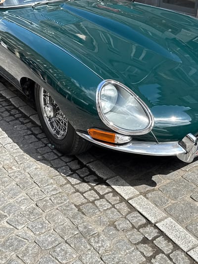 A close-up, eye-level shot captures the front-left quadrant of a vintage, dark green Jaguar E-Type parked on a sun-dappled cobblestone street. The car's highly reflective paintwork mirrors the surrounding architecture, including building facades and windows, suggesting an urban setting. The polished chrome of the headlight bezel and bumper gleams, contrasting with the orange turn signal. The intricate wire-spoke wheel is visible, peeking out from beneath the fender. The cobblestone pavement, a characteristic feature of Bruges, Belgium, is laid in an irregular pattern, with patches of sunlight and shadow indicating a bright, possibly midday, scene. No people are present in the frame.
