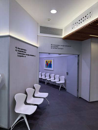 This is a wide-angle, eye-level, indoor photograph of a modern clinic waiting area in Palma de Mallorca, Spain. The image captures a sterile, minimalist environment with a focus on white furniture and muted grey walls.
In the foreground, three white, molded plastic chairs with thin metal legs are arranged against a grey textured wall. The chairs have a distinctive curved back and seat. The floor is covered with large, dark grey tiles that have a subtle white veining pattern.
On the left wall, there are two sets of directional signs with black lettering and symbols. The upper sign, pointing left, reads "Àrea quirúrgica" (Surgical Area). Below it, also pointing left, is "Hospitalització" (Hospitalization). To the right of these, a sign points forward and reads "Endoscòpies i cirurgia ambulatòria" (Endoscopies and ambulatory surgery). Below this, another sign points forward and reads "Ecografies" (Ultrasounds).
On the right side of the image, an open doorway leads into another room. Above the doorway, a similar set of directional signs are mounted on the wall, pointing upwards: "Endoscòpies i cirurgia ambulatoria", "Ecografies", and "Admissió quirúrgica" (Surgical Admission). Inside the room visible through the doorway, there are more white chairs and a framed piece of colorful abstract art on the wall.
The ceiling features recessed lighting and ventilation grilles. A distinct wooden slatted panel runs along the upper right portion of the ceiling. The overall impression is one of cleanliness, efficiency, and a modern healthcare facility. There are no people visible in the photograph.
