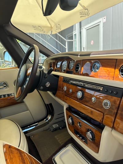 The image captures the interior of a luxurious Rolls-Royce car parked outdoors in Dub, Montenegro. The car features a cream-colored leather interior with extensive polished wood trim, particularly on the dashboard and center console. The dashboard is adorned with classic circular gauges and air vents. A detailed shot of the steering wheel reveals its leather and wood construction. The focus is on the opulent dashboard area, showcasing intricate controls and a central infotainment system. The exterior environment is visible through the car's windshield, hinting at a sunny day and a building with a metal facade. The overall impression is one of high-end automotive luxury juxtaposed with a modest outdoor setting.