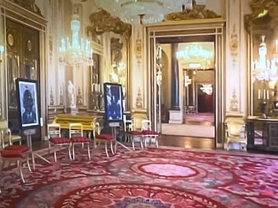 The image depicts a grand, ornate room with high ceilings adorned with chandeliers and intricate gold detailing. The walls are a pale color with decorative trim. A large, plush red carpet with a complex floral pattern covers the floor. In the foreground, several small, red-cushioned stools with delicate gold legs are arranged. To the left, a tall, framed black and white portrait of a person is displayed on a stand. In the mid-ground, a bright yellow, low table or bench is visible, with more chairs arranged around it. Further into the room, a doorway opens into a hallway, also richly decorated with chandeliers and mirrored surfaces, creating a sense of depth. A dark wooden cabinet with glass doors is positioned to the far right. There are no people visible in the image. The scene suggests a formal setting, possibly a museum, gallery, or a historic building. There is no discernible information within the image that places the location in Liverpool, United Kingdom.
