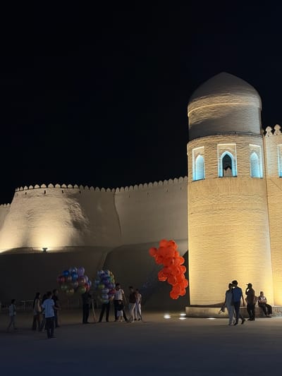 The image depicts a nighttime scene in the historic city of Khiva, Uzbekistan, likely within the walled inner city of Itchan Kala, near one of its prominent towers. In the foreground, a group of people are gathered. To the left, several individuals are standing and conversing, with one person holding a large bunch of colorful balloons. To the right, another person is selling a cluster of red balloons. In the mid-ground, a couple walks hand-in-hand, while other individuals are also present, some sitting and others standing.

Dominating the right side of the frame is a tall, cylindrical tower constructed of light-colored brick, illuminated by artificial lighting that accentuates its ribbed texture. The tower features arched windows with blue light emanating from within. Adjacent to this tower, on the left, is a substantial white wall, characteristic of Khiva's ancient fortifications, also illuminated from below. The wall is topped with crenellations, and there are small openings visible along its length. The background is a dark night sky. The overall atmosphere is one of a lively evening gathering against a backdrop of ancient architecture.