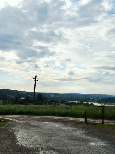 The image depicts a cloudy sky over a rural landscape in Bolshaya Usa, Russia. In the foreground, a wet, gravelly dirt road curves into the scene, with puddles reflecting the overcast sky. To the right, a rusty metal fence with chain-link infill encloses an area of tall green grass. Beyond the road and fence, a line of dense green trees and vegetation stretches across the middle ground, with scattered buildings and structures visible amongst them. A wooden utility pole with insulators stands prominently in the left-middle ground, supporting electrical wires that extend across the frame. In the distance, rolling hills covered in trees form the horizon, with a body of water, possibly a river or lake, visible in the center distance. The overall atmosphere is muted and somewhat damp, suggesting recent rain. There are no people or animals visible in the image.