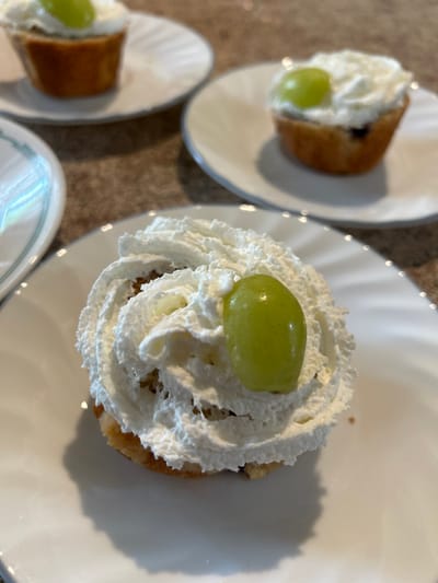 The image displays an indoor scene featuring three individual desserts arranged on a light brown, textured surface, likely a countertop. The primary subject is a dessert positioned in the foreground, centered and in sharp focus. It consists of a golden-brown, baked item, resembling a muffin or cupcake base, generously topped with white whipped cream. A single, oval-shaped, bright green grape rests on the whipped cream. This dessert sits on a plain white, circular plate.
In the midground and background, two additional similar desserts are visible, out of focus. One to the upper right consists of a golden-brown baked item, possibly containing dark specks like blueberries, topped with white whipped cream and two green grapes. Another partially visible dessert to the upper left also shows a baked base, whipped cream, and a single green grape, all on identical white plates. The lighting is soft and even. No individuals or specific identifying features of a larger setting within Thousand Oaks, United States, are present; the scene depicts food on a domestic surface.
