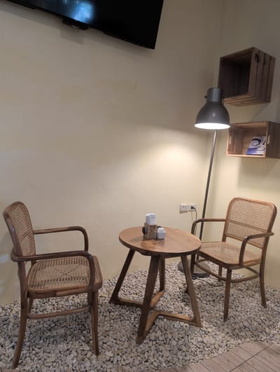 This interior scene features a small seating arrangement positioned on a floor covered in small, white pebbles. The arrangement consists of two wooden chairs with woven backs and seats, flanking a small, round wooden table. On the table, there are items that appear to be disposable coffee cups or containers.
A floor lamp with a gray, dome-shaped head is positioned behind the table and chairs, providing illumination. The scene is set against a light beige wall. Wall-mounted wooden crates are positioned higher on the wall. The environment appears to be an indoor space, and the style suggests a cafe or lounge. Based on the prompt, the location is identified as Thong Sala, Thailand, but there is no direct evidence of this in the image. A television screen is visible on the left.