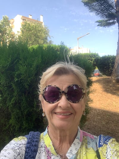 This is a selfie taken outdoors in Odivelas, Portugal, likely during the daytime given the bright sunlight and clear blue sky. The subject is a woman with blonde hair, wearing large, round sunglasses with a leopard-print frame. She is smiling, and her face is turned towards the camera. She is wearing a colorful, patterned shirt with blue, yellow, and pink elements. A navy blue, textured accessory, possibly a bag or scarf, is visible on her right side.
The background consists of lush green foliage, including dense bushes and trees, and a patch of dry grass and dirt. In the distance, a building with a cream-colored facade and chimneys can be seen, along with a street light and a hint of a palm tree on the left side of the frame. The overall impression is of a sunny day in a suburban or park-like setting. The image is oriented vertically, as if the camera was held in portrait mode.