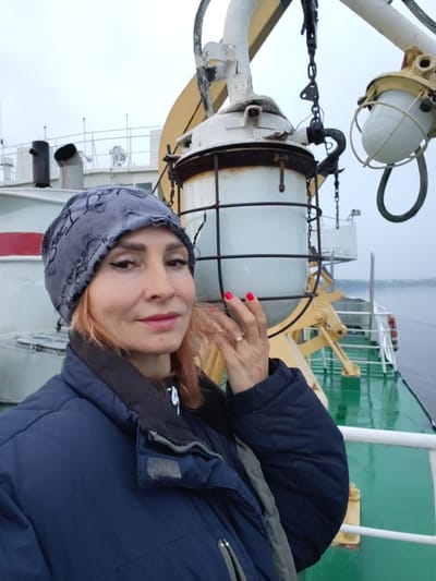 A woman with blonde hair and a gray beanie takes a selfie on a boat. She is wearing a dark blue jacket and her fingernails are painted red. Behind her, part of the boat's structure is visible, including metal railings, a yellow support beam, and a light fixture. The water is visible in the background. The scene is likely outdoors, possibly on a ferry or a ship.