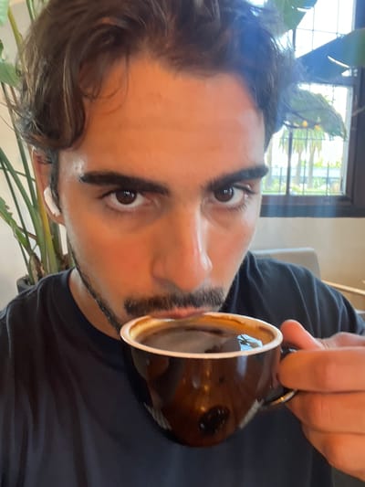 A man is holding a coffee cup close to his mouth, as if about to drink. He is wearing a dark t-shirt and has dark hair and eyebrows. A window with plants is behind him, indicating he may be indoors. The location in the city of Avellaneda, Argentina, is not discernable from the image.