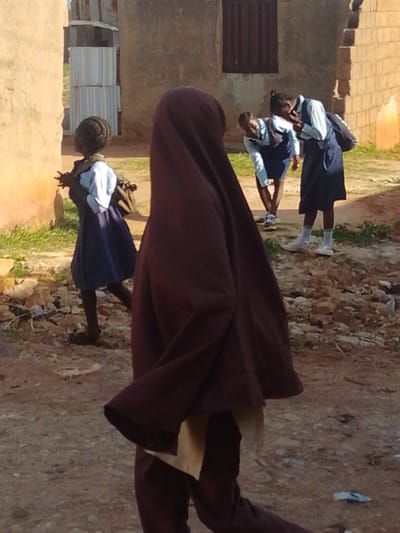 In Tumfure, Nigeria, four young individuals are captured in a moment outdoors. A person in the foreground is seen from behind, wearing a dark maroon headscarf and matching trousers, with a light-colored fabric peeking out from underneath. To their left, a child in a blue dress and white shirt is walking, with their right arm raised and hand slightly open. Further back, two other children, likely students, are interacting. One, on the right, wears a dark blue dress over a white shirt and carries a dark backpack. The other, to their left, is bent over, possibly speaking to the first. The environment is dusty and unpaved, with low-lying green vegetation and scattered rocks and debris. In the background, a building with a textured wall and a dark-framed window stands, alongside a structure made of corrugated metal sheets. The lighting suggests it is daytime, with sunlight illuminating the scene.