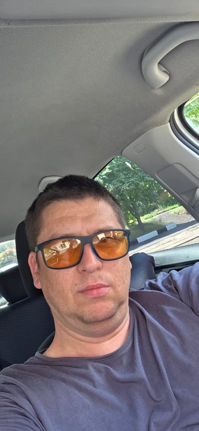 The media file shows a selfie of a man inside a car, with the image rotated 90 degrees counter-clockwise. The man is wearing a dark t-shirt and dark-rimmed sunglasses with orange lenses. His face is partially visible, with his mouth closed and his gaze directed slightly upwards and to the left. The background inside the car shows the gray interior of the car's ceiling and a white handle. Through the car window, trees and greenery are visible, suggesting an outdoor setting. The lighting indicates it is daytime. The image is grainy and appears to be taken with a mobile phone. No visible text is present. The context provided indicates the location is Donetsk, Ukraine.