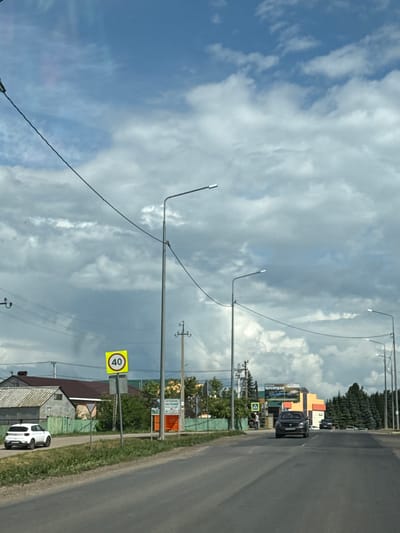This image depicts a street scene in Nurykeevo, Russia. A paved road runs from the foreground towards the background, bordered by grassy verges and a green wooden fence on the left. Several streetlights are visible along the road, with overhead wires connecting them. A white SUV is parked on the left side of the road. Further down the road on the left, there are some residential buildings with pitched roofs. A yellow speed limit sign indicating "40" is mounted on a pole. On the right side of the road, a colorful building with signs is visible, and a line of coniferous trees stands behind it. Two cars are driving on the road in the distance. The sky is partially cloudy.
