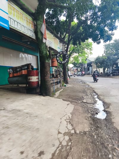 The image shows a street scene in Baleendah, Indonesia, taken from a perspective looking down the road. It is likely taken from a vehicle in motion, judging by the blur of the pavement.  A large tree with a thick trunk is at the left of the image, partially obscuring a business storefront. An Indonesian flag hangs from the building, with a red and white banner, likely advertising the business, obscuring the shop's name. A red cylindrical container is visible at the base of the tree. The road is cracked and shows signs of wear and tear, with a small puddle of water reflecting the sky. There are a few vehicles in the distance, including a motorcycle. The street appears to be a minor residential road, with a few houses or businesses visible in the background.  The foliage is dense, indicating a tropical climate. The scene suggests a somewhat neglected or rundown area, with a focus on local businesses and limited traffic.