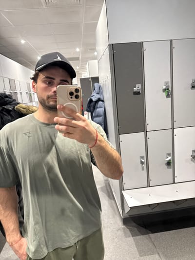 A male individual is depicted taking a mirror selfie inside what appears to be a gym changing room or locker room in Coventry, United Kingdom. The subject, a male estimated to be between 20 and 35 years of age, has short dark hair visible under a black baseball cap featuring a white Jumpman logo. He has facial stubble and is wearing a faded olive green short-sleeve t-shirt with a dark Gymshark logo on the left chest, and olive green sweatpants. A thin red string bracelet is visible on his left wrist. He holds a gold-colored smartphone, equipped with a clear case and a white circular MagSafe ring, in his right hand to capture the image.
The background, visible in the mirror's reflection, consists of multiple banks of light grey and dark grey metal lockers, each fitted with key locks and numerical labels (e.g., "27", "9"). Below the lockers, a white bench or seating unit is present. Several items of clothing, including a dark blue puffer jacket and another jacket with a fur-lined hood, are seen hanging on hooks within the locker area. The room is illuminated by overhead recessed lighting. The floor is a light grey, consistent with industrial or gym flooring.