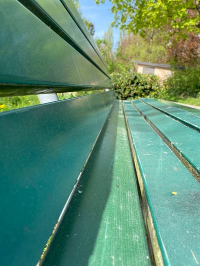 The image, rotated approximately 90 degrees counter-clockwise, presents a close-up, low-angle view of a vibrant green park bench bathed in bright sunlight. The bench is composed of multiple parallel wooden slats, painted a deep, slightly worn green. The perspective emphasizes the length of the bench, with the slats extending from the foreground into the upper right background, creating strong diagonal leading lines.

The surface of the wooden slats exhibits signs of use, including minor scratches, scuffs, and areas where the paint appears subtly faded or chipped. Small, dark specks and a tiny yellow flake are visible on the surfaces, particularly on the slats closer to the viewer. Gaps between the individual wooden planks reveal darker, possibly weathered wood or underlying structure. Glimpses of metal supports are visible where the slats meet their framework.

The background is softly blurred but clearly depicts an outdoor, natural setting. Lush green foliage from trees and bushes dominates the mid-ground, interspersed with areas of reddish-brown leaves, suggesting a mix of deciduous trees. A clear, bright blue sky is visible above the trees on the left side of the frame. A light-colored building or structure can be faintly seen through the foliage in the distant background.

The strong contrast between illuminated and shadowed areas on the bench indicates a sunny day with clear weather, likely during the morning or afternoon. The bench is empty, with no people or animals present, and no particular activity is taking place. There is no visible text within the image. The scene is set in Lucerne, Switzerland.