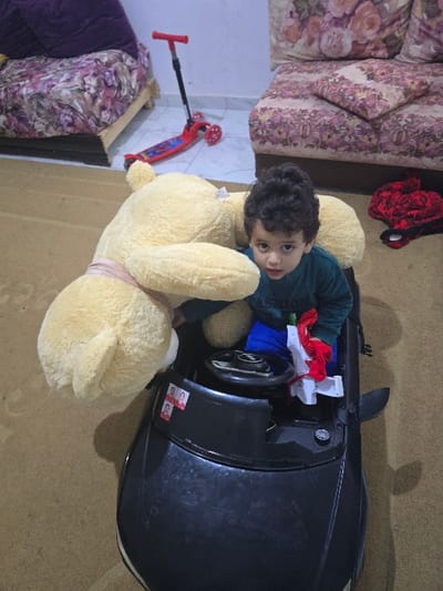 A young child sits in a black toy car, cradled by a large, light yellow teddy bear. The child is wearing a dark green shirt. The car is filled with red and white objects. A red scooter lies on the floor nearby. A floral couch is visible in the background, along with a purple blanket on a different couch. The floor appears to be a light tan color.