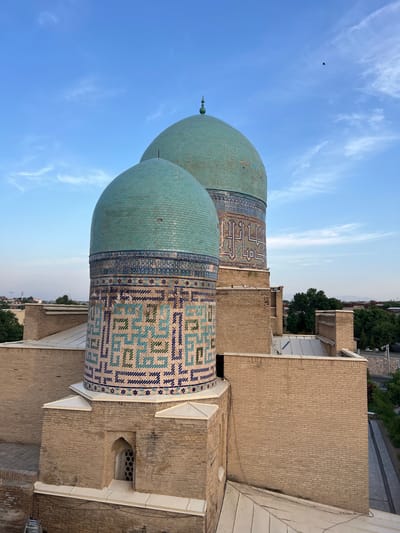 The image showcases two turquoise domes of a building, likely a mosque or mausoleum, in Samarkand, Uzbekistan. The domes are adorned with intricate geometric patterns in blue and white tiles. The building itself is constructed from light-brown bricks. The sky above is a clear blue with wispy white clouds, suggesting a pleasant day. A single bird is visible in the sky. The perspective is from a higher vantage point, looking down at the domes and surrounding buildings. Trees and more brick structures are visible in the background, indicating an urban or historical complex. The time of day appears to be late afternoon or early evening, given the soft light and the color of the sky. The image conveys a sense of architectural grandeur and historical significance. There is no visible text in the image.