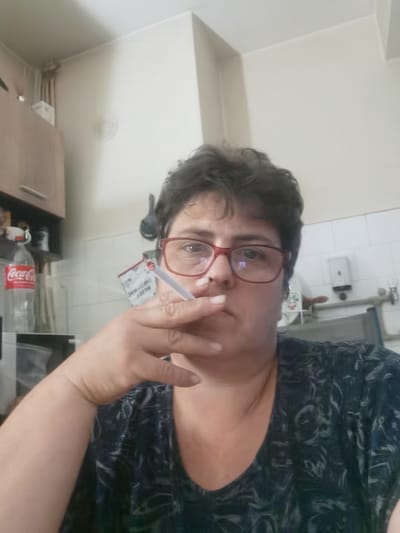 An indoor, medium-shot, selfie-style photograph shows a person with short, dark, curly hair and red-framed glasses. The person is holding a lit cigarette in their right hand, near their mouth, with smoke obscuring part of their face. They are wearing a dark blue t-shirt with a light grey, swirling pattern. In the background, a white tiled wall is visible, with some kitchen cabinets to the left. A bottle of Coca-Cola is on a shelf in the cabinets. The environment appears to be a kitchen or similar domestic setting. The location is not explicitly identifiable as Lomets, Bulgaria, based solely on the visual information present.