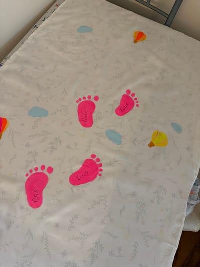 A white mattress pad with a floral design rests on a mattress and frame. Scattered across the pad are cartoonish depictions of bare footprints, colored hot pink and each labeled with a number in handwritten text: "one", "two", "three", and "four." Also on the pad are small, stylized drawings of hot air balloons, colored yellow/brown and orange/brown, along with three small blue shapes that could represent raindrops or blobs. The scene's location is unspecified and unidentifiable in Bearpark, United Kingdom.