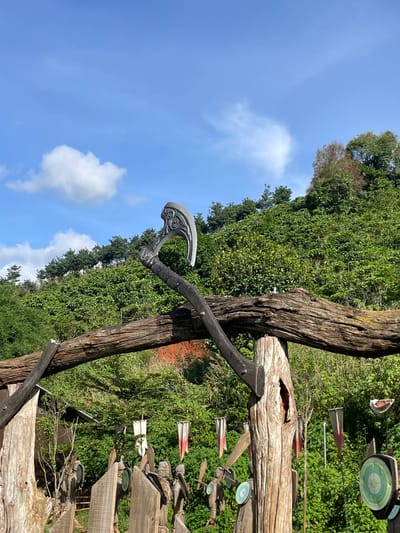 An elaborate, prop axe rests on a weathered wooden archway in Thôn 1, Vietnam. The axe, with a decorative, crescent-shaped blade, is propped on a thick, gnarled log that forms the horizontal beam of the arch. The arch itself is constructed from rough, unfinished logs. Behind the arch, a lush, green hillside is densely covered in vegetation, with a clear blue sky overhead dotted with a few white clouds. Below the arch, wooden figures that resemble warriors, some adorned with flags and circular shields, stand in a line. The scene appears to be an outdoor display or attraction.