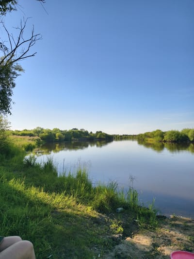 A serene, wide-angle landscape photograph captures a tranquil river scene under a clear, vast blue sky. The river, calm and reflective, mirrors the lush green foliage of trees and bushes lining its banks, creating a symmetrical, peaceful composition. The sun, positioned to the left of the frame, casts a warm glow on the verdant grass and sandy shore in the foreground. On the left bank, a person's legs are visible, seated and partially cropped out of the frame, suggesting a moment of rest or observation. The overall atmosphere is one of natural beauty and quietude, likely during the daytime. The location is identified as Nikolskoye, Russia, a setting that evokes a sense of rural or suburban tranquility.