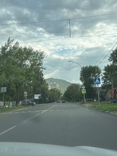 The image shows a view from a vehicle traveling on a paved road towards a mountainous landscape under a cloudy sky. The road has white lane markings and a pedestrian crossing. On the left side of the road, there are trees, a dark SUV parked, and a billboard. On the right side, there are more trees, a truck loaded with hay, and a residential building in the background. Traffic signs are visible, including a pedestrian crossing sign and a speed bump warning sign. The mountain in the distance has a rocky, uneven surface. The overall impression is that of a semi-urban or suburban environment on the outskirts of a city. The location is identified as Mineralnye Vody, Russia, a city known for its proximity to the Caucasus Mountains.