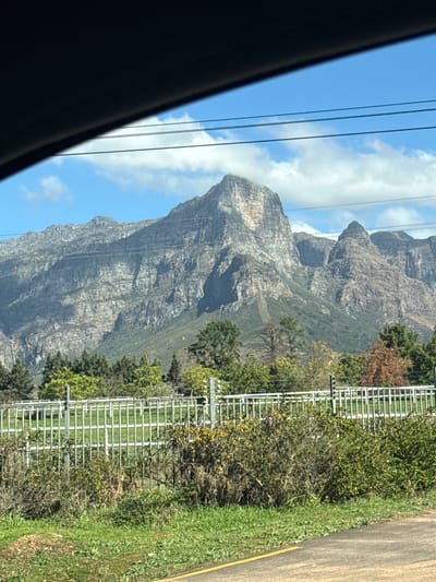 A landscape view from a car window features a massive, rocky mountain with visible stratifications. Power lines run parallel to the mountain's slope, extending from the top left. To the right of the mountain is a white, grid-like fence, adjacent to a grassy area and a portion of a road marked with a yellow line. A variety of trees and bushes are interspersed between the fence and the mountain. The sky is partly cloudy. The location is Stellenbosch Nu, South Africa.