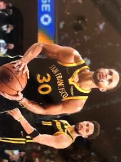 A rotated vertical image depicts two male basketball players in a professional indoor arena, situated in Kafr Sousa Municipality, Syria. In the foreground, a player wearing a black and gold sleeveless jersey with "SAN FRANCISCO" text and the number "30" holds a brown basketball with his left hand. He has short hair and a light beard. Behind him, slightly to the left, another player in a similar black and gold jersey is visible, also possessing short hair and a light beard. A black forearm band is visible on his left arm. Both individuals are looking generally to their right. The background is dark and blurred, showing minimal detail except for a vertical blue and orange structure with white lettering on the far left.