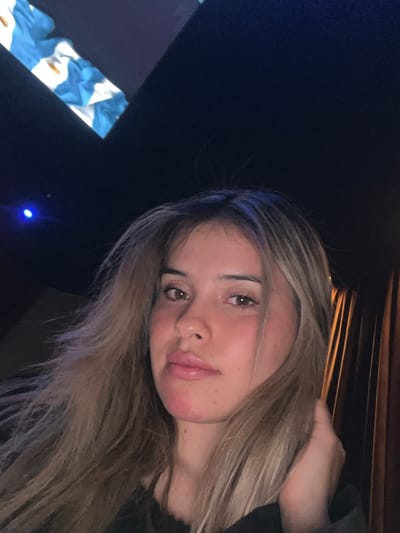 A young woman with blonde hair is captured in a close-up selfie. She is looking directly at the camera, with her hair loosely framing her face. The background is dark, with hints of blue light on the left and vertical lines of warm-toned fabric on the right. The overall impression is of an indoor setting, possibly a dimly lit room or a vehicle. The image does not contain any information about the city of San Fernando del Valle de Catamarca, Argentina.