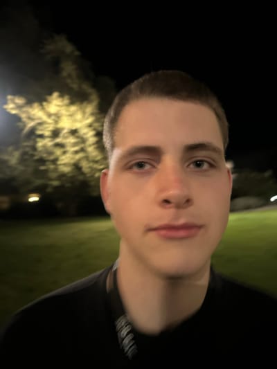 The image displays a close-up, frontal portrait of a young male, appearing to be Caucasian, with short, dark hair styled in a buzz cut and light-colored eyes. He wears a dark-colored shirt and a lanyard with white text or symbols around his neck. A faint reddish mark is visible on the bridge of his nose. The subject's gaze is directed towards the camera, and his expression is neutral. The scene is set outdoors at night in Guimarães, Portugal. The background is out of focus, revealing indistinct green ground cover, likely grass, and blurred yellow light sources, suggesting illuminated trees or foliage, consistent with street lighting.