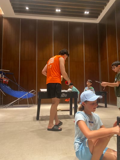 The image shows an indoor scene with at least five individuals, possibly in a recreation or activity area. The backdrop consists of a dark wood panelled wall with metallic horizontal accents.

Foreground: A male figure wearing an orange t-shirt and black shorts is holding a red object, possibly a weight or ball. He is positioned beside a black metal structure with the word "Elite" visible on its side. Beside him is a small, partially collapsed blue portable basketball hoop.

Midground: To the right, a person wearing a light blue shirt and a blue cap is seated at a black table. There are three additional individuals further in the background, engaged in various activities. One appears to be holding a string-like object.

General Environment: The floor appears to be carpeted. The lighting is bright and even. The overall scene suggests a casual setting where various activities are taking place.

Based on visual cues, there's nothing to suggest that the image was taken in Marina Maroma, Mexico.