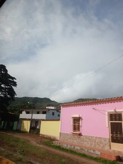 This is a medium-shot, outdoor, daytime photograph of a residential street in Tacarigua, Venezuela. The foreground shows a dirt road with dry grass and some green vegetation. To the right, a pink-colored house with a tiled roof is visible. This house features a window with decorative iron bars and a beige door with vertical bars. The lower part of the pink house is adorned with stone-like tiles. Behind the pink house, a yellow wall and a white building with a tiled roof are partially visible. This white building appears to have multiple windows. Further in the background, lush green mountains are shrouded in clouds. The sky is mostly cloudy with patches of blue. A dark-leaved tree is present on the left side of the image. A thin electrical wire stretches diagonally across the upper portion of the frame. There are no people or animals visible in the scene.