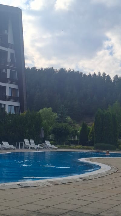 The image shows an outdoor swimming pool with a person partially submerged in the blue water. The pool is surrounded by a stone patio and white lounge chairs. In the background, there is a dense forest with lush green trees. A modern building with several windows is visible to the left of the pool. The sky is overcast with some clouds, suggesting a daytime scene. The overall impression is one of a tranquil resort or hotel setting, possibly in a mountainous or forested area, given the location context of Razlog, Bulgaria. The lighting indicates it is likely daytime, though the cloud cover may be obscuring direct sunlight. There is no visible text in the image.
