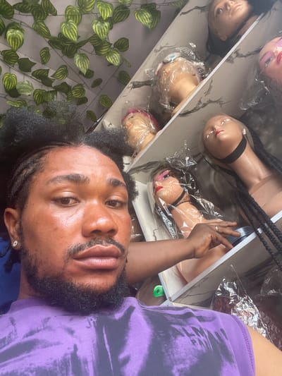 A man is taking a selfie in what appears to be a wig shop, identifiable by the mannequin heads displayed on shelves behind him. The shop wall is decorated with artificial vines. The location appears to be Alaiyabiagba, Nigeria, although the image itself provides no direct evidence to confirm this. The man is wearing a purple shirt. The mannequin heads are covered in plastic wrap, some with wigs or braided hairstyles.