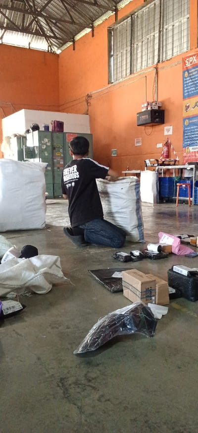 A young male, appearing to be in his late teens or early twenties, kneels on a concrete floor within a warehouse. He is wearing a black t-shirt with white stripes on the sleeves and the text "FOREVER UNDERDOGS" on the back, along with blue jeans and black shoes. He is reaching into a large, white woven bag that is partially filled with what appears to be material or goods. The warehouse environment is characterized by an orange-painted wall, a corrugated metal roof supported by exposed beams, and fluorescent lighting.

In the background, there are metal lockers, a white counter with items on it, and shelving units holding blue plastic crates and other supplies. A fire extinguisher is visible on a stand. The floor is littered with various packages, including cardboard boxes, some wrapped in plastic, and other items that are difficult to identify precisely due to the image quality and distance. A large white bag is positioned to the left of the kneeling male, and another similar bag is to his right. The scene suggests an active workspace, likely involved in packaging or sorting goods for distribution. The overall impression is that of a functional, albeit somewhat cluttered, industrial or storage facility. The location is identified as Kiangroke, Indonesia.