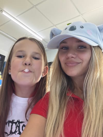 Two females are depicted in a selfie, with the foreground occupied by a younger girl with brown hair and a white t-shirt, her mouth open with a piece of white bubble gum mid-pop. To her right, a blonde female wears a light blue cap with fuzzy koala ears and a pink embroidered koala face, along with a red shirt. Both are indoors, suggested by the fluorescent lighting and tiled ceiling visible in the background. The location of Thep Krasatti, Thailand, is not determinable from the visual information provided.