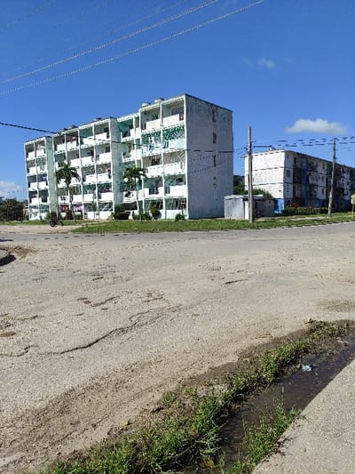 This image depicts a residential scene in Ciego de Ávila, Cuba, under a clear blue sky with sparse clouds. The foreground features a weathered, light-colored asphalt or concrete road surface, exhibiting numerous cracks and areas of erosion. Along the lower right edge, a narrow, unpaved ditch or gutter runs parallel to the road, containing standing water and a strip of green grass and weeds.
In the midground, two multi-story apartment buildings are visible. The prominent building on the left is a six-story structure, appearing light green or off-white, with visible signs of weathering and discoloration. Each floor features multiple balconies, many of which have clothes hanging to dry. Two small palm trees stand in front of this building at ground level. To the right of the first building, a utility pole with multiple overhead wires extends into the sky. A small, light-colored utility box or shed is positioned behind this pole. Further right and more distant, a second, smaller, multi-story building, also exhibiting signs of wear and neglect, is partially visible. Additional utility wires stretch across the upper portion of the frame. No individuals are visible in the scene, though the hanging laundry indicates human habitation. A bicycle is partially visible near the base of the prominent building on the left.