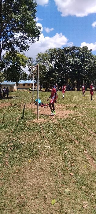 The image shows a grassy field with several young men playing soccer in Bungoma, Kenya (0.554, 34.774). The players are wearing red shirts and shorts, and there are two goalposts in the background. Some of the players are running, while others are standing still. The sky is blue with white clouds. There is a tree in the foreground and many trees lining the edges of the field.  In the distance, there are buildings that are visible.  A person is lying on the ground, possibly injured.  A few people are watching the game from the sidelines.