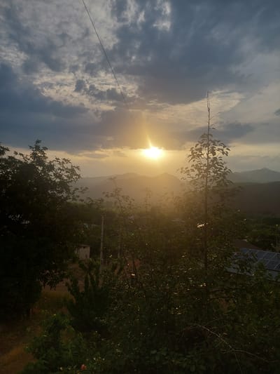 This is a photograph taken outdoors in the city of Areni, Armenia, during a sunset. The sun is visible in the upper center of the frame, partially obscured by clouds and trees, casting a warm glow across the sky. The sky is filled with dramatic, heavy clouds, some dark and others lighter with the sun's rays breaking through. In the foreground, there are several trees with lush green foliage, some of which are silhouetted against the bright sky. The landscape behind the trees appears to be hilly or mountainous, with layers of fading mountains visible in the distance. A power line stretches diagonally across the upper part of the image. In the mid-ground, there are glimpses of buildings, including a small white structure and what appears to be a roof with solar panels. The overall impression is a serene, natural scene with a touch of human habitation.
