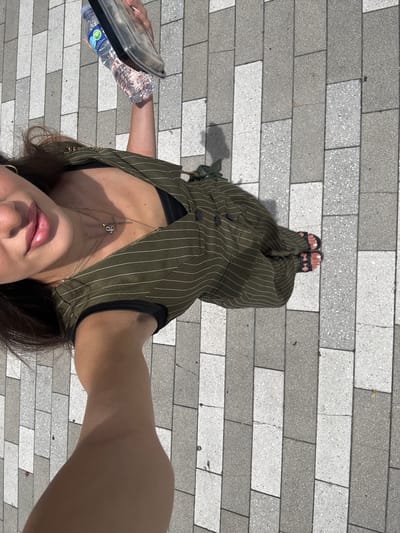An overhead, high-angle full shot captures a person from the chest down, holding a clear plastic water bottle and a clear plastic container. The person is wearing a green pinstripe jumpsuit, a black bra, and black sandals. Their left arm is extended towards the camera. The ground is paved with rectangular grey and white tiles. The image is cropped to focus on the person and the immediate surroundings. The scene appears to be outdoors in an urban setting, likely a public walkway.