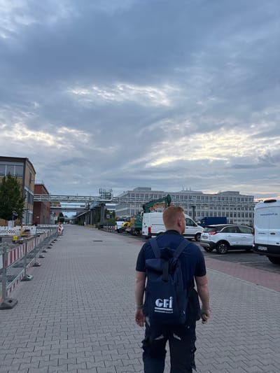 The image captures a person from behind, walking away from the camera on a paved walkway lined with construction barriers. The person is wearing a dark blue t-shirt, dark pants, and a dark blue backpack with a white logo that reads "GFI GESELLSCHAFT FUR INDUSTRIETECHNIK mBH & CO. KG". Several vehicles, including white vans and a white car, are parked on the right side of the walkway. In the background, large industrial buildings and overhead piping systems are visible under a cloudy sky. The scene appears to be an industrial or commercial area. Based on the presence of a white van with "www.kohlhoff.de" written on it, and the vehicle registration plate starting with "MA", which is the standard German license plate code for the city of Mannheim, the location is identified as Mannheim, Germany.
