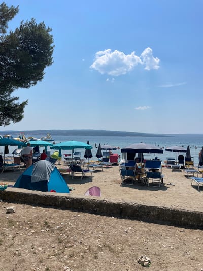 The image depicts a crowded beach scene in Crikvenica, Croatia. The orientation of the photo is vertical, presenting a view of the shoreline from right to left. Numerous beach chairs and umbrellas are set up on the sandy beach, with many people relaxing or wading in the clear blue water.
In the foreground, a low stone wall separates the sandy beach from a rougher, rocky area. Several beach chairs, some occupied, are visible along the sand. Further back, rows of blue and white striped beach umbrellas provide shade, with people scattered beneath them. The sea stretches out to the horizon, where a faint outline of hills can be seen. The sky is a bright, clear blue with a few fluffy white clouds, suggesting a sunny day.
The scene appears to be during daylight hours, likely mid-morning or afternoon, given the bright sunlight and shadows. The atmosphere is one of leisure and recreation, typical of a summer beach day. The presence of many people suggests a popular tourist destination. The overall impression is a lively and inviting beach environment. No discernible text is present in the image.