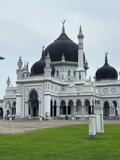 The image shows a grand mosque with multiple domes and minarets, located in Alor Setar, Malaysia. The mosque is predominantly white with dark domes. It features intricate architectural details, including arched walkways, decorative patterns, and crescent moon finials on its spires. In the foreground, a well-maintained lawn leads up to the mosque's entrance, which is flanked by pillars and a wide set of stairs. Two individuals are visible sitting on a bench in the middle ground, near the entrance. The sky is overcast, suggesting a cloudy day.