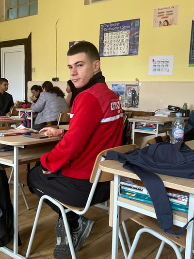 The image depicts an indoor classroom scene in Cherven Bryag, Bulgaria. A young male individual, identified by a jacket bearing "ЧЕРВЕН БРЯГ," is positioned in the foreground, seated at a light wood-topped desk with a white metal frame, looking directly towards the camera. He has short, dark hair and wears a red long-sleeved jacket with white horizontal stripes and a black collar, black athletic pants, and dark athletic shoes with prominent soles. His left hand rests on a tablet device on his desk, and a red and white bracelet is visible on his left wrist.

In the mid-ground, other students are seated at similar desks. To the left of the main subject, another male is partially visible, wearing a dark top, facing away from the camera. Further back, a female student in a grey jacket is seen looking downwards. The classroom features pale yellow walls and light-colored wooden floorboards. Educational posters are affixed to the wall, including one titled "The Teacher Development Series" displaying phonetic symbols, and another titled "Peaceful Life." A water bottle labeled "Балдаран" is visible on a desk in the mid-ground to the right. In the immediate right foreground, a navy blue backpack lies on another desk, beneath which several books and notebooks are stored on a shelf. A white door with a dark frame and an upper window pane are visible in the background on the left.