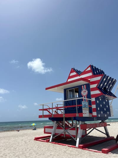 This is a vertical, eye-level, full shot of a lifeguard tower on Miami Beach. The tower is shaped like a star and is painted with red and white stripes and blue with white stars, reminiscent of the American flag. The tower has a red and white railing around a small platform. Text on the tower reads "MIAMI BEACH" and "8 ST". The background shows a calm ocean with gentle waves, a sandy beach, and a bright blue sky with scattered clouds. In the distance, two people are sitting under a yellow and blue umbrella on the beach. The overall scene conveys a patriotic and serene beach atmosphere.