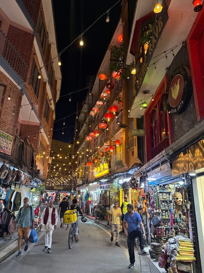 Captured in Kathmandu Metropolitan City, Nepal, the image depicts a bustling street scene at dusk or night. The perspective is that of standing amidst the crowd, looking down the street.
Overhead, strings of warm-toned lights and red lanterns crisscross, providing illumination. Architectural styles vary, with brick buildings on one side and structures adorned with decorative elements like red accents and signage on the other.
The street is filled with people walking in both directions. Visible among them are men wearing traditional clothing like kurtas and other outfits indicative of the local culture. A cyclist wearing a yellow delivery bag is also seen.
On either side of the street, shops display their wares, including clothing and various goods, enhancing the atmosphere of a lively market. Signs like "Gothic Rave Bar Estd. 2075" can be seen.
Overall, the image encapsulates the vibrant nightlife and urban life of Kathmandu, Nepal, with its blend of cultural elements and commercial activity.