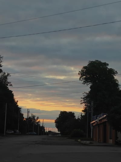 The image depicts a street scene at dusk in Hadiach, Ukraine. The sky is filled with overcast clouds, tinged with the orange and yellow hues of a setting sun. Several power lines crisscross the sky. On the left side of the road, a line of trees partially obscures parked cars. On the right, a small, modern-looking building with a segmented roofline stands next to a utility pole. The road itself appears to be paved but worn, with some potholes visible. The overall atmosphere is subdued due to the low light conditions.