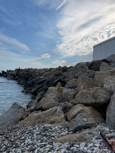 The photograph shows a view of a rocky breakwater in Olginka, Russia, on a day with a partially cloudy sky. The breakwater is constructed from large, irregularly shaped gray and brown rocks. The sea can be seen to the left of the breakwater, and a small strip of shore covered in smooth, gray pebbles is visible on the right. Several people are standing along the breakwater, some appear to be fishing with fishing rods. A white rectangular structure is visible at the top left of the breakwater. A hand is holding a black phone over the rock on the breakwater.